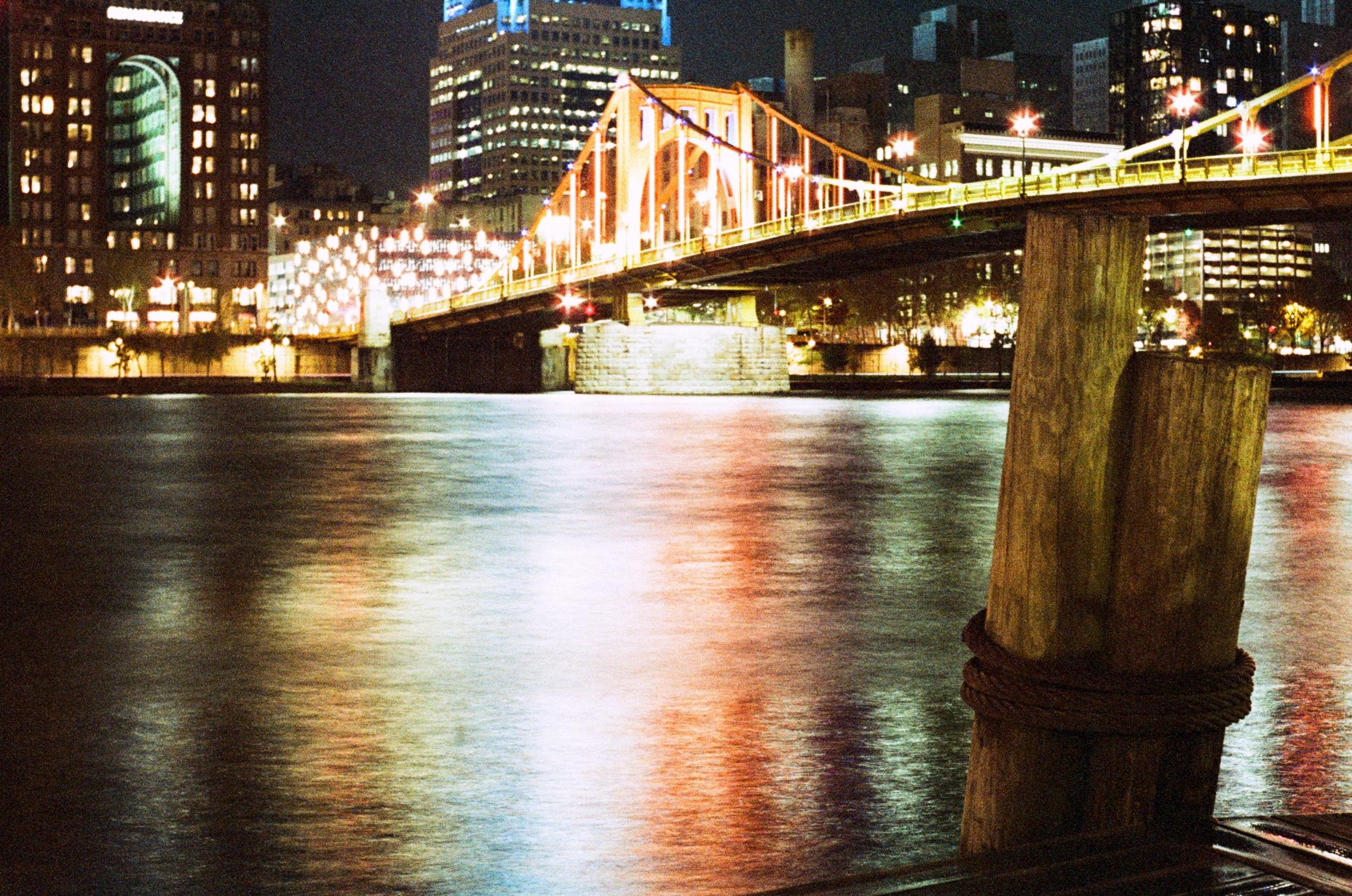 color photo, bright red and yellow bridge in the background, foreground’s got a lit up docking stake for boats to tie to. in between some really colorful allegheny river water.
